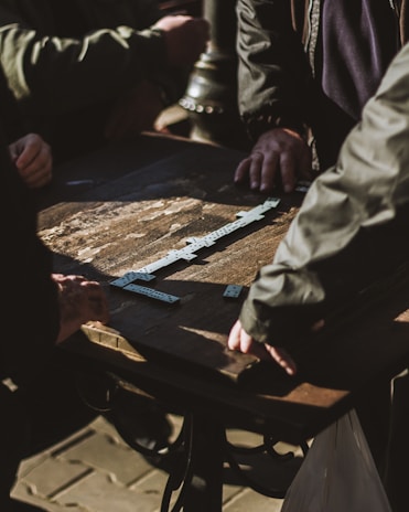 Several individuals are gathered around a wooden table engaging in a game of dominoes. The sunlight casts shadows across the table, highlighting the domino pieces meticulously arranged in play.