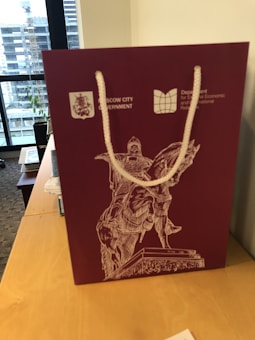A maroon gift bag with white rope handles. The bag features a detailed drawing of an equestrian statue and logos for the Moscow City Government and Department for External Economic and International Relations. It is placed on a wooden table near a window with buildings visible outside.