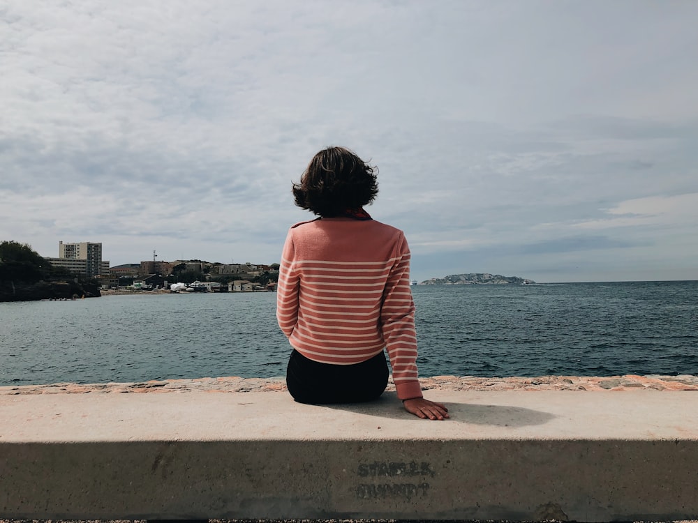 woman sitting on concrete pavement overlooking body of water during daytime