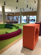 A modern indoor space with a colorful seating area featuring circular benches in green and purple colors. The room has a wooden floor and is well-lit with overhead lighting. Large windows and bookshelves line the walls, and a few people are visible near the entrance.