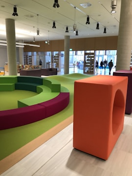 A modern indoor space with a colorful seating area featuring circular benches in green and purple colors. The room has a wooden floor and is well-lit with overhead lighting. Large windows and bookshelves line the walls, and a few people are visible near the entrance.