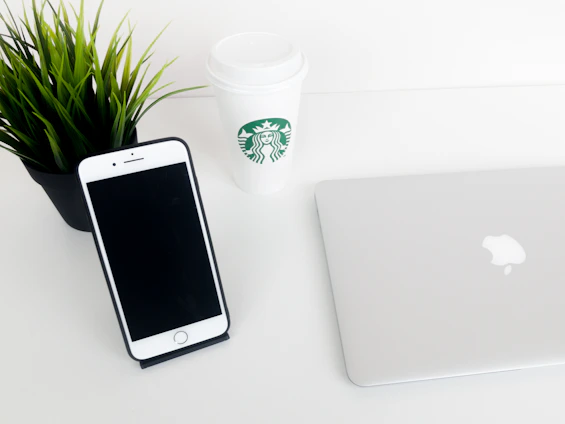 A cozy home desk setup featuring a sleek stabiliq phone stand holding a smartphone next to a laptop and a cup of coffee.