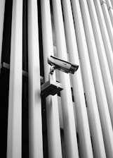 A security camera is mounted on a wall with vertical metallic bars, creating a sense of surveillance and modern architecture. The linear pattern of the bars contributes to the industrial and minimalist aesthetic of the scene.