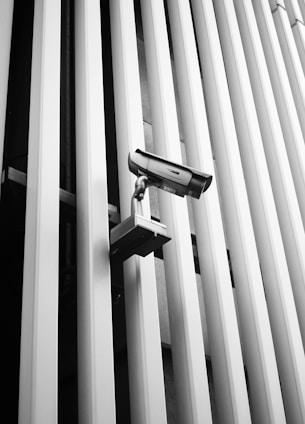 A security camera is mounted on a wall with vertical metallic bars, creating a sense of surveillance and modern architecture. The linear pattern of the bars contributes to the industrial and minimalist aesthetic of the scene.