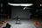 A dark room featuring a professional photography or film studio setup. There is a single black folding chair positioned in the middle of a gray backdrop. Various pieces of equipment such as boom microphones, lighting stands, and sandbags are visible. An exit sign is illuminated in the background.