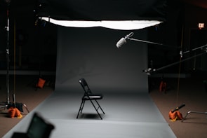 A dark room featuring a professional photography or film studio setup. There is a single black folding chair positioned in the middle of a gray backdrop. Various pieces of equipment such as boom microphones, lighting stands, and sandbags are visible. An exit sign is illuminated in the background.