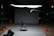 A dark room featuring a professional photography or film studio setup. There is a single black folding chair positioned in the middle of a gray backdrop. Various pieces of equipment such as boom microphones, lighting stands, and sandbags are visible. An exit sign is illuminated in the background.