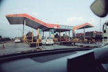 A toll plaza with multiple lanes for vehicles, featuring a distinctive red roof and various signs. Several vehicles, including cars and a truck, are passing through, and traffic cones are placed to guide the traffic. The surrounding area includes some buildings and a clear sky above.