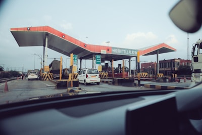 A toll plaza with multiple lanes for vehicles, featuring a distinctive red roof and various signs. Several vehicles, including cars and a truck, are passing through, and traffic cones are placed to guide the traffic. The surrounding area includes some buildings and a clear sky above.