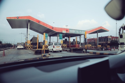 A toll plaza with multiple lanes for vehicles, featuring a distinctive red roof and various signs. Several vehicles, including cars and a truck, are passing through, and traffic cones are placed to guide the traffic. The surrounding area includes some buildings and a clear sky above.