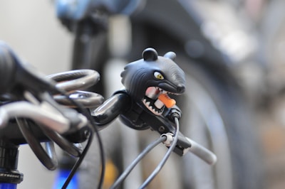 A quirky bicycle bell shaped like a bear with an open mouth and protruding tongue is attached to the handlebars. The background is blurred, focusing attention on the bell and a coiled cable lock wrapped around the handlebar.