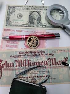 Various types of currency are displayed, including a US one-dollar bill, a Zimbabwean trillion-dollar note, and a historical German note for ten million marks. A magnifying glass, a red pen, and a novelty Bitcoin coin are also present.