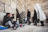 A man is sitting on the ground with a camera and tripod, intently capturing a photograph. He is positioned next to a stone wall with an archway. Nearby, a person in traditional attire with a white shawl and black stripes walks by, while another person in a black jacket and cap looks in the direction of the camera.