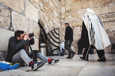A man is sitting on the ground with a camera and tripod, intently capturing a photograph. He is positioned next to a stone wall with an archway. Nearby, a person in traditional attire with a white shawl and black stripes walks by, while another person in a black jacket and cap looks in the direction of the camera.