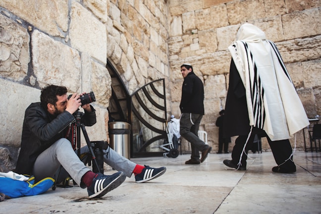 A man is sitting on the ground with a camera and tripod, intently capturing a photograph. He is positioned next to a stone wall with an archway. Nearby, a person in traditional attire with a white shawl and black stripes walks by, while another person in a black jacket and cap looks in the direction of the camera.