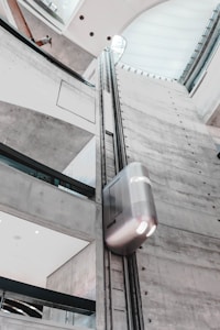 A modern indoor elevator ascends along a tall, gray cement shaft within a building. The architecture features clean lines and large windows allowing natural light to stream in. The elevator itself has a sleek, metallic finish.