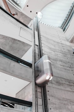 A modern indoor elevator ascends along a tall, gray cement shaft within a building. The architecture features clean lines and large windows allowing natural light to stream in. The elevator itself has a sleek, metallic finish.