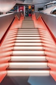 A staircase with illuminated steps and red side walls leads to an upper level. Above the stairs, there are framed sports memorabilia on display. To the right, part of a car is visible, hinting at an automobile-themed area.