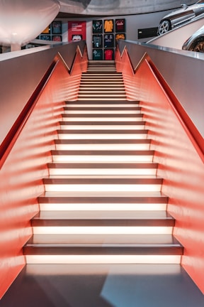 A staircase with illuminated steps and red side walls leads to an upper level. Above the stairs, there are framed sports memorabilia on display. To the right, part of a car is visible, hinting at an automobile-themed area.