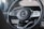 A close-up of a car steering wheel featuring a central logo with surrounding control buttons. The steering wheel is made from a combination of materials including perforated leather and metallic elements.