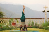 A person is practicing yoga on a blue mat in a garden setting. They are in a headstand position, wearing green leggings and a tank top. The garden is adorned with various plants and flowers, and a decorative fence and hanging pots are visible. In the background, there are misty hills and overcast skies.
