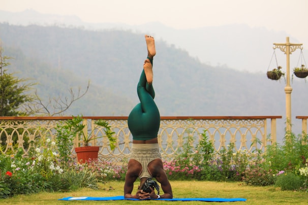 A person is practicing yoga on a blue mat in a garden setting. They are in a headstand position, wearing green leggings and a tank top. The garden is adorned with various plants and flowers, and a decorative fence and hanging pots are visible. In the background, there are misty hills and overcast skies.
