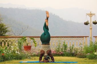 A person is practicing yoga on a blue mat in a garden setting. They are in a headstand position, wearing green leggings and a tank top. The garden is adorned with various plants and flowers, and a decorative fence and hanging pots are visible. In the background, there are misty hills and overcast skies.