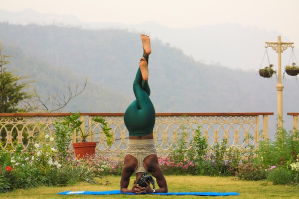 A person is practicing yoga on a blue mat in a garden setting. They are in a headstand position, wearing green leggings and a tank top. The garden is adorned with various plants and flowers, and a decorative fence and hanging pots are visible. In the background, there are misty hills and overcast skies.