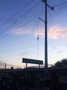 A street sign displays 'QUERETARO 77' beneath a clear sky during sunset. The scene includes silhouettes of people in the foreground and electrical poles with wires extending across the frame. A building and some trees are visible in the background.