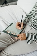 A person wearing a striped shirt is holding a pen and writing on a clipboard with paper. The person is seated on a bed or sofa with white sheets, and only part of the torso and legs are visible.
