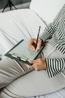 A person wearing a striped shirt is holding a pen and writing on a clipboard with paper. The person is seated on a bed or sofa with white sheets, and only part of the torso and legs are visible.