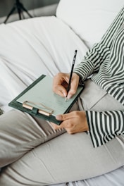 A person wearing a striped shirt is holding a pen and writing on a clipboard with paper. The person is seated on a bed or sofa with white sheets, and only part of the torso and legs are visible.