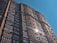 A tall, modern building with numerous windows and air conditioning units mounted on the walls. The sun reflects off the windows, creating a bright glare. The structure has a mix of dark red and brown bricks, and it stretches high into a clear blue sky.