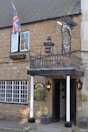 A rustic brick building features a sign for 'Ilchester Hotel' under a traditional inn-style sign with a crest. A Union Jack flag is prominently displayed next to an open window. The entrance is framed by two white pillars, flanked by outdoor lanterns, and potted shrubs. The windows have white frames, adding contrast to the light brown stone facade.