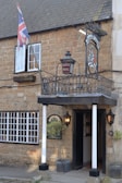 A rustic brick building features a sign for 'Ilchester Hotel' under a traditional inn-style sign with a crest. A Union Jack flag is prominently displayed next to an open window. The entrance is framed by two white pillars, flanked by outdoor lanterns, and potted shrubs. The windows have white frames, adding contrast to the light brown stone facade.