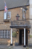 A rustic brick building features a sign for 'Ilchester Hotel' under a traditional inn-style sign with a crest. A Union Jack flag is prominently displayed next to an open window. The entrance is framed by two white pillars, flanked by outdoor lanterns, and potted shrubs. The windows have white frames, adding contrast to the light brown stone facade.