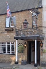 A rustic brick building features a sign for 'Ilchester Hotel' under a traditional inn-style sign with a crest. A Union Jack flag is prominently displayed next to an open window. The entrance is framed by two white pillars, flanked by outdoor lanterns, and potted shrubs. The windows have white frames, adding contrast to the light brown stone facade.