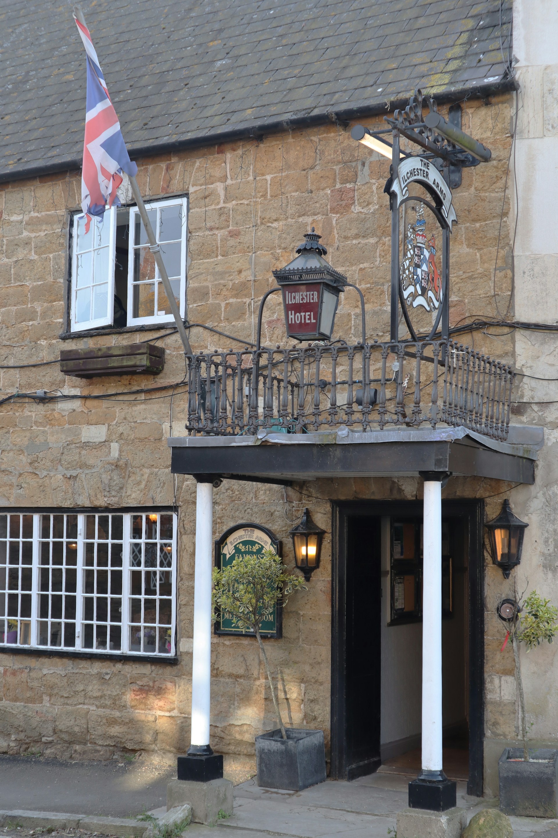 A rustic brick building features a sign for 'Ilchester Hotel' under a traditional inn-style sign with a crest. A Union Jack flag is prominently displayed next to an open window. The entrance is framed by two white pillars, flanked by outdoor lanterns, and potted shrubs. The windows have white frames, adding contrast to the light brown stone facade.