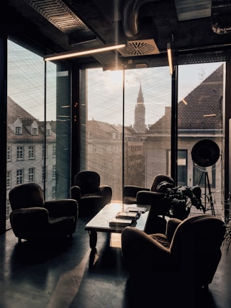 A modern office or lounge area with large floor-to-ceiling windows offering a view of city buildings and a distant spire. The room features several cushioned armchairs arranged around a low coffee table with books and a magazine. There is a large, industrial-style lamp in the corner, and a plant adds a touch of greenery to the space. The lighting is subdued, creating a cozy atmosphere.