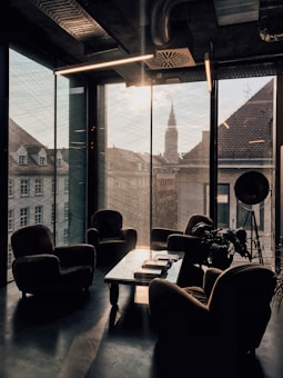 A modern office or lounge area with large floor-to-ceiling windows offering a view of city buildings and a distant spire. The room features several cushioned armchairs arranged around a low coffee table with books and a magazine. There is a large, industrial-style lamp in the corner, and a plant adds a touch of greenery to the space. The lighting is subdued, creating a cozy atmosphere.