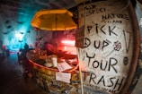 A dimly lit, urban bar with a gritty, underground vibe. A large yellow umbrella hangs over a cluttered counter filled with cups and trays. The walls are covered in graffiti, including messages promoting environmental responsibility like 'Pick up your trash.' The background features a bluish illuminated area with people and silhouettes of birds in flight.
