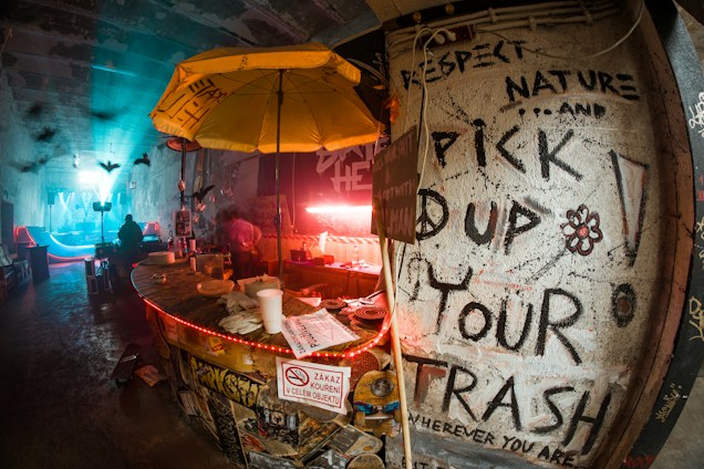 A dimly lit, urban bar with a gritty, underground vibe. A large yellow umbrella hangs over a cluttered counter filled with cups and trays. The walls are covered in graffiti, including messages promoting environmental responsibility like 'Pick up your trash.' The background features a bluish illuminated area with people and silhouettes of birds in flight.