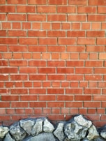 A brick wall with a pattern of horizontal and vertical lines is topped by a row of unevenly shaped, textured stones at the base. The bricks are a reddish-brown color with visible grout lines.