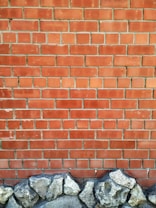 A brick wall with a pattern of horizontal and vertical lines is topped by a row of unevenly shaped, textured stones at the base. The bricks are a reddish-brown color with visible grout lines.