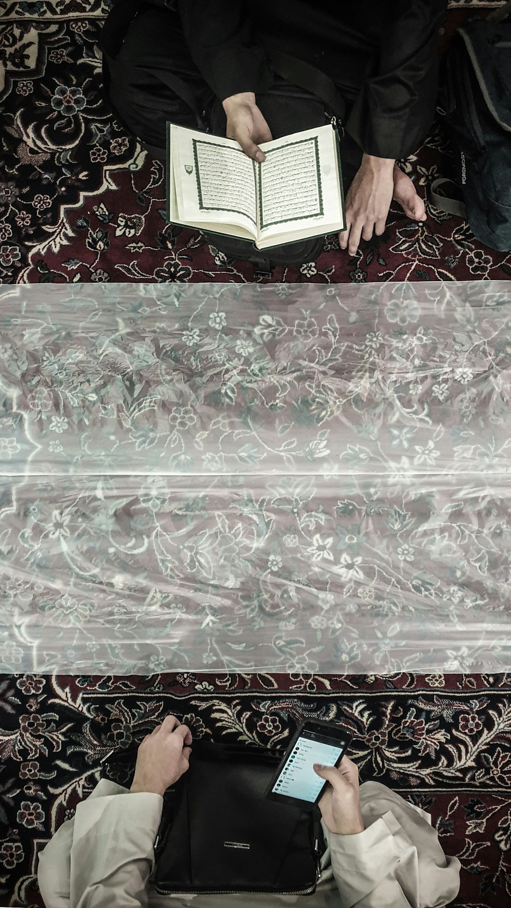zwei Personen, die auf einem Teppich sitzen, eine mit dem Koran und die andere mit Smartphone