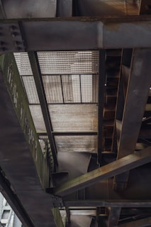 A close-up view of the underside of a metallic structure with a girded, grid-like pattern visible. The structure displays a combination of grey and dark green hues, with intricate steel beams intersecting each other. The image showcases a complex arrangement of geometric lines and industrial design.