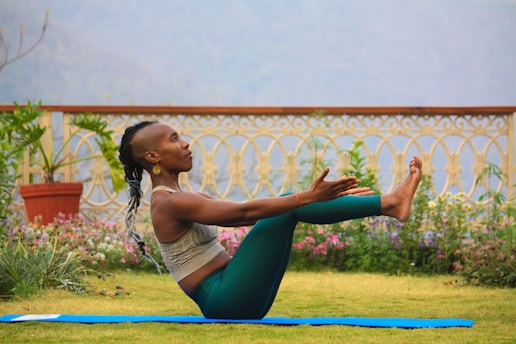 A person is practicing yoga outdoors on a grassy area, balanced in a boat pose with arms extended forward and legs raised. The setting includes a decorative railing with intricate patterns and a backdrop of flower beds. The atmosphere appears tranquil and focused.