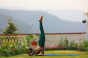 A person performs a yoga pose on a blue mat in a garden setting, with lush greenery and a decorative fence in the background. Beyond the garden, misty mountains are visible under a cloudy sky, creating a serene environment.