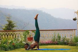 A person performs a yoga pose on a blue mat in a garden setting, with lush greenery and a decorative fence in the background. Beyond the garden, misty mountains are visible under a cloudy sky, creating a serene environment.