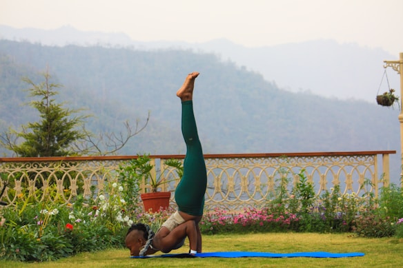 A person performs a yoga pose on a blue mat in a garden setting, with lush greenery and a decorative fence in the background. Beyond the garden, misty mountains are visible under a cloudy sky, creating a serene environment.
